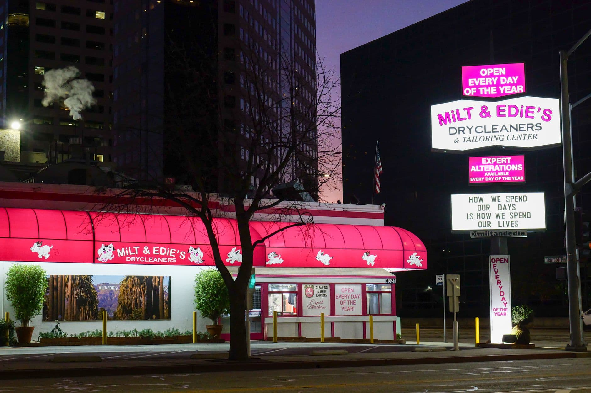 Milt & Edie's Drycleaners illuminated at night in bustling USA cityscape.