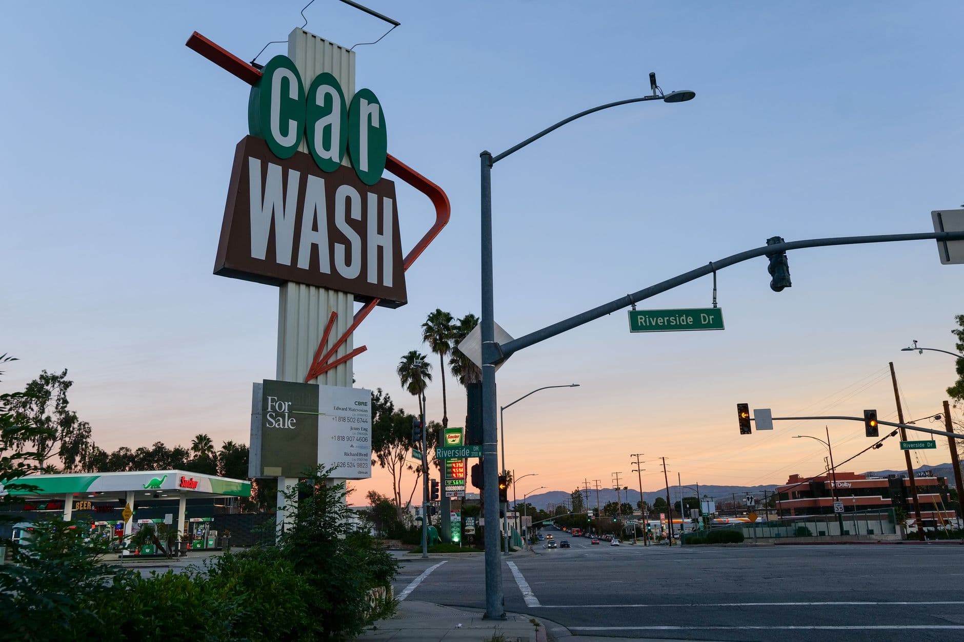 A retro car wash sign at sunset on Riverside Drive with a gas station in the background.