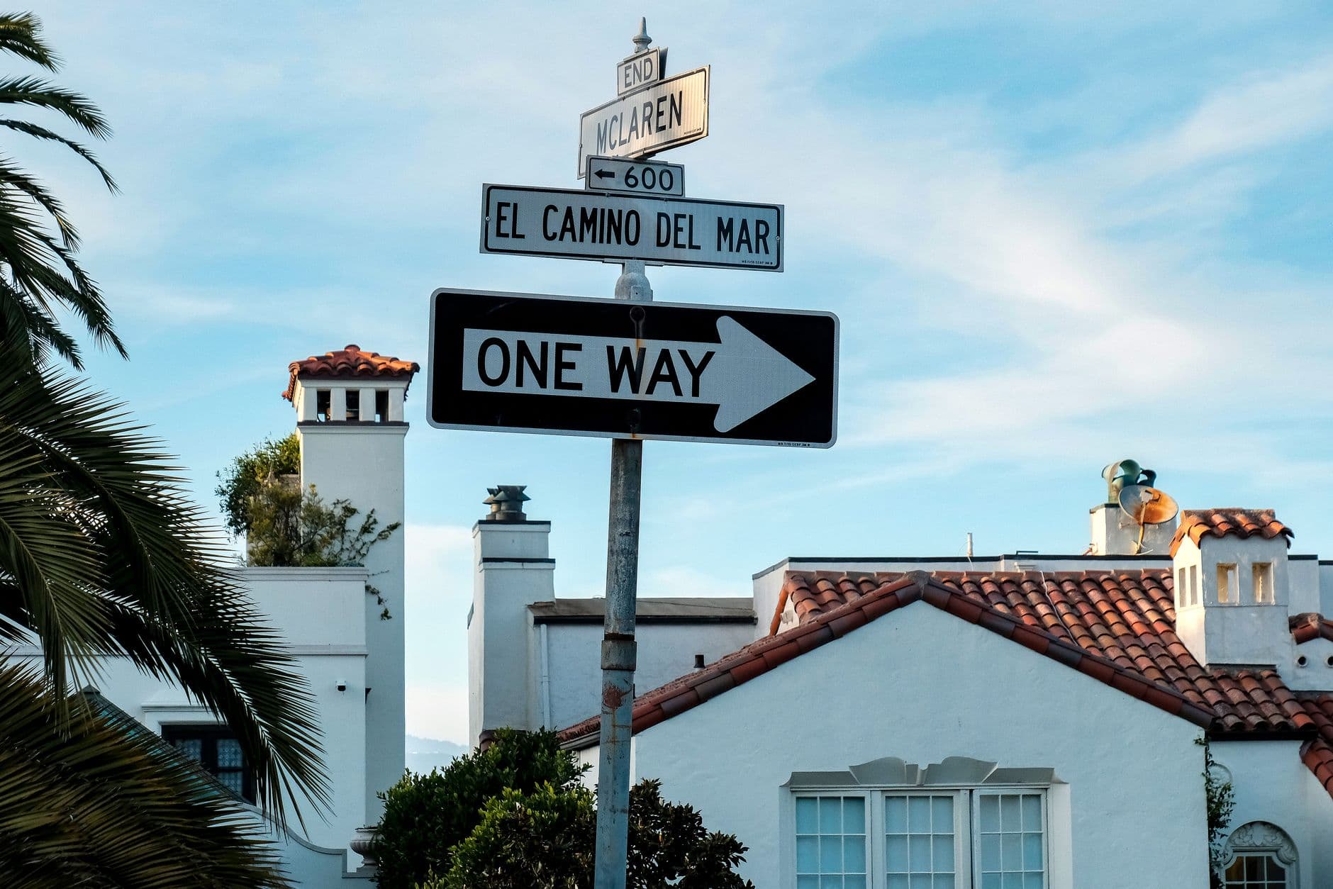 Street signs with Spanish-style architecture, depicting direction and local street names on a sunny day.