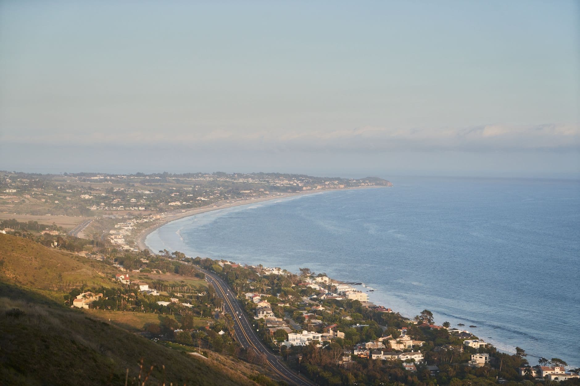 Distance view of calm sea and coastal settlement with asphalt road running through green hilly terrain in foggy weather