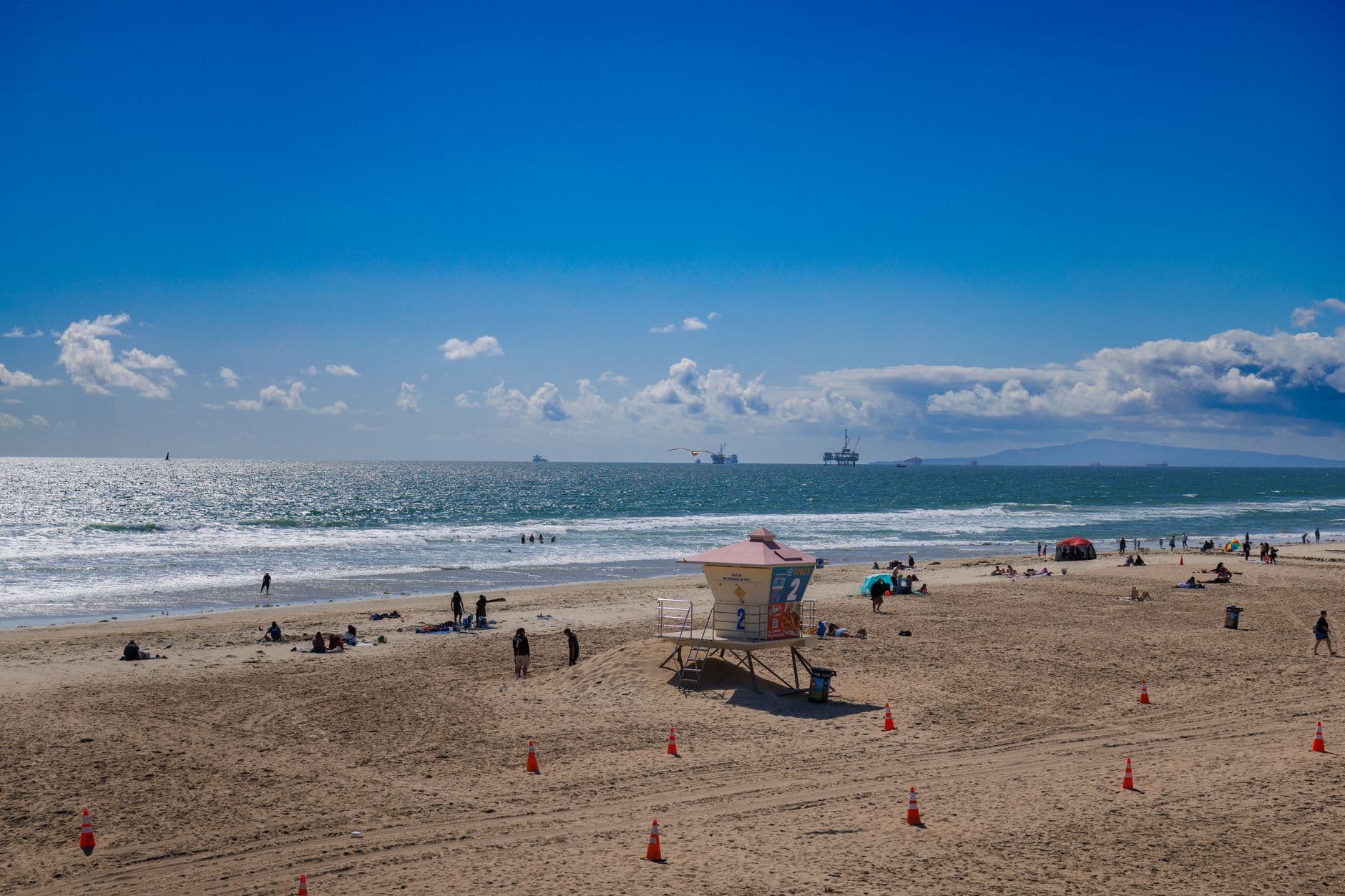 Panoramic view of a sunny day at Huntington Beach, featuring a lifeguard tower, people enjoying the beach, and ships on the horizon.