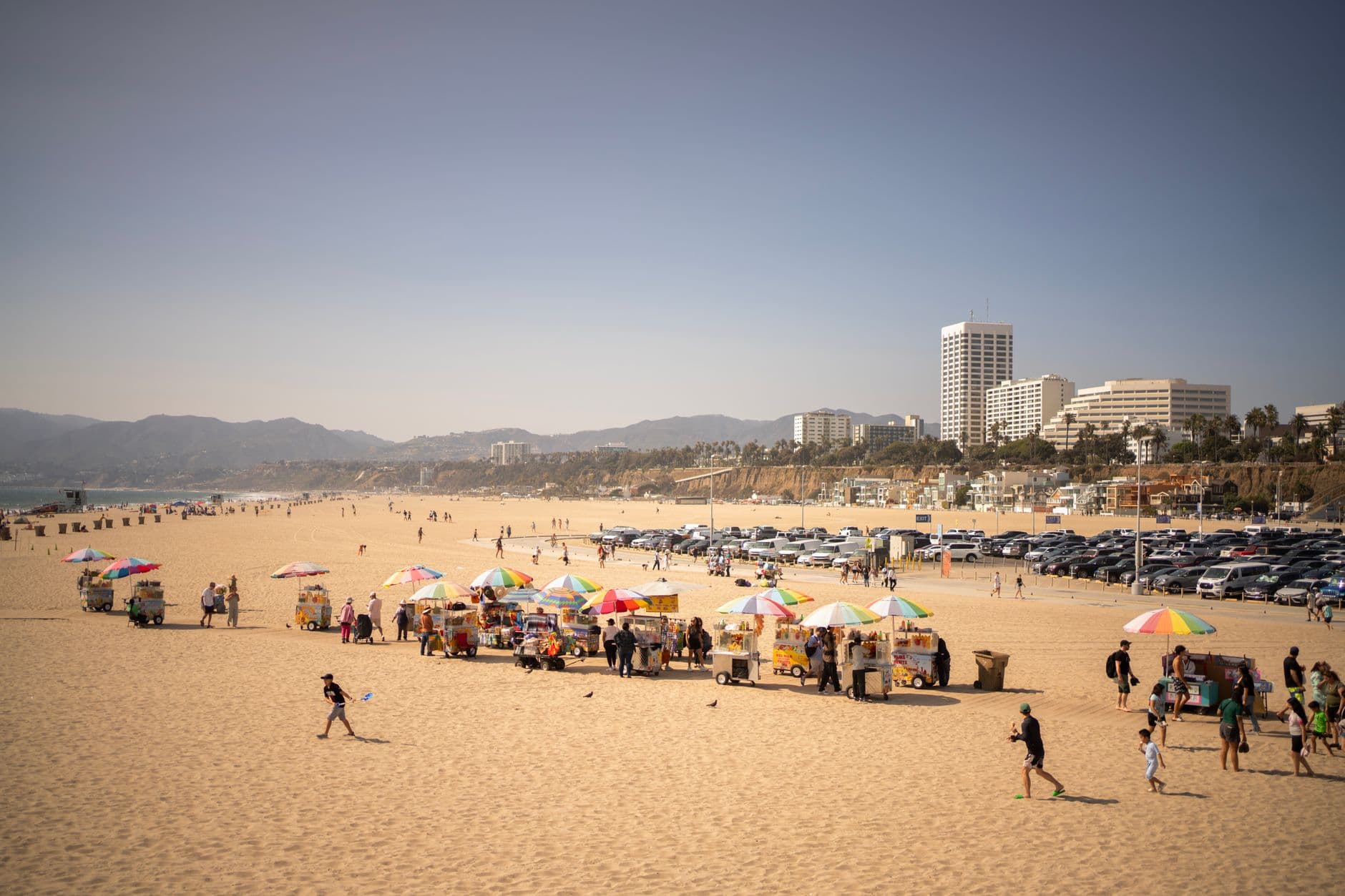 Sunny day at Santa Monica Beach with colorful vendor stalls and people strolling the sandy shore.